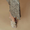 A close-up of a person's foot wearing fuzzy beige double-strap sandals with gold buckle details, paired with a leopard-print skirt, against a solid beige background.