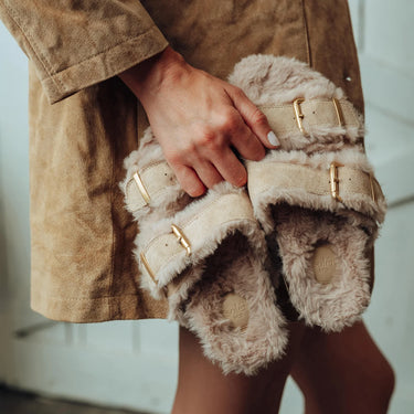 A person in a brown suede jacket holds a pair of beige faux-fur slide sandals with gold-tone buckle straps. The sandals are shown close up, with the person's hand and legs visible against a light indoor background.