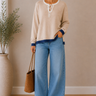 Woman wearing a beige sweater and blue jeans standing indoors with a plant and bag in the background.