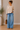 Woman wearing a beige sweater and blue jeans standing indoors with a plant and bag in the background.