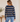 A woman shown from the back wearing a navy blue and beige horizontally striped long-sleeve knit sweater with light blue jeans, standing in an indoor setting.