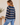 A woman wearing a navy blue and beige striped long-sleeve sweater, paired with light blue jeans, shown from a side-back angle in a softly lit indoor setting.