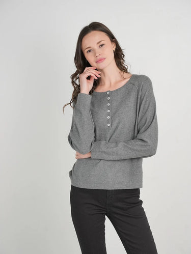 A woman modeling a gray long-sleeve knit top with a buttoned neckline, paired with black pants, against a plain light gray studio background.