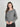 A woman modeling a heather gray long-sleeve henley top with a button placket, paired with black pants, against a light gray studio background.