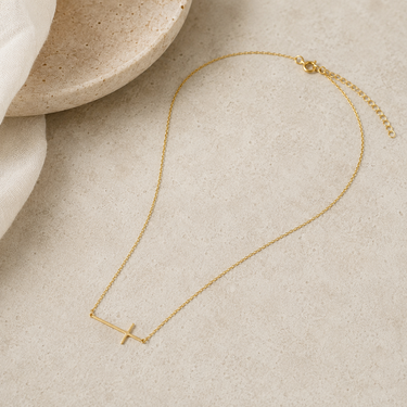 A delicate gold chain necklace with a minimalist bar pendant in the shape of a cross, displayed on a textured beige surface with a neutral stone dish and white fabric in the background.