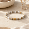 A slim beaded bracelet with alternating white and gold ceramic beads, displayed on a textured beige stone surface with a ceramic bowl and dried flowers in the background.
