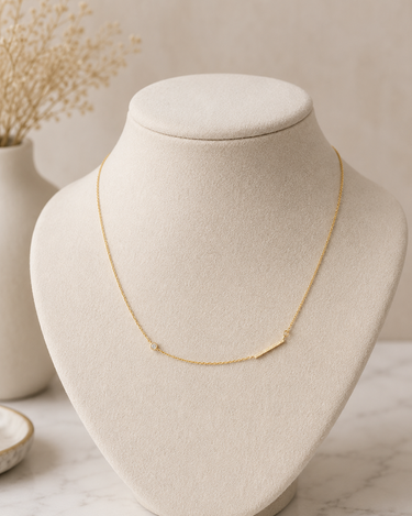 A delicate gold necklace with a minimalist bar design, displayed on a cream-colored jewelry bust against a soft beige background.