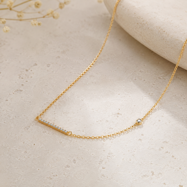A delicate gold chain necklace with a bar-shaped pavé setting and a small round accent stone, displayed on a textured light beige surface.