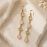 A pair of gold drop earrings with peach-pink and light green gemstone beads and small clear stones, displayed on a textured beige stone surface with soft fabric and dried flowers.