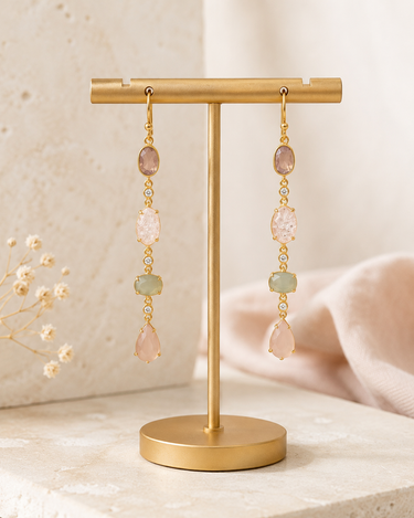 A pair of gold drop earrings with pink, peach, and light green gemstones, displayed on a gold stand against a soft beige background.