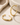 A pair of polished gold-tone hoop earrings displayed on a light beige textured surface with soft neutral props in the background.