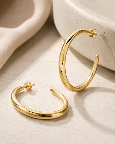 A pair of polished gold-tone hoop earrings displayed on a light beige textured surface with soft neutral props in the background.