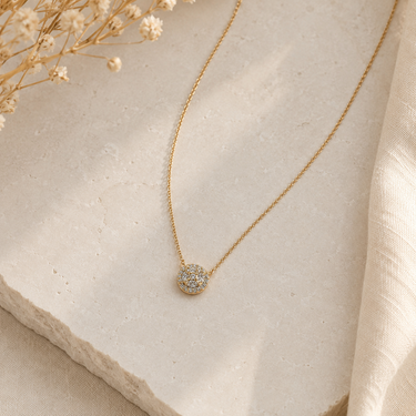 A delicate gold chain necklace with a small, round pavé-set pendant, displayed on a textured light beige stone slab with dried baby's breath flowers.