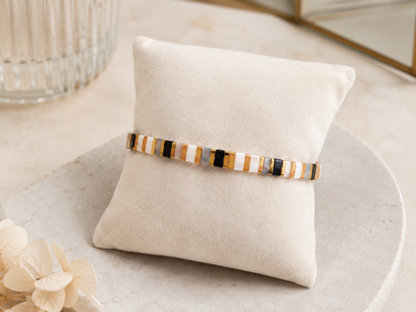 A beaded stretch bracelet with alternating white, gold, and black rectangular beads, displayed on a cream-colored velvet jewelry pillow on a light marble surface.