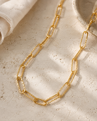 A gold hammered paper clip necklace on a textured surface.