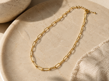 A gold paperclip chain necklace displayed on a round beige stone tray with a textured fabric napkin in the corner.