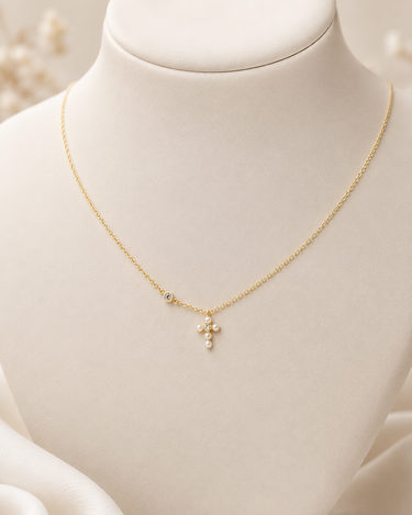 A delicate gold chain necklace with a small cross pendant made of tiny white pearls, displayed on a cream-colored jewelry bust.