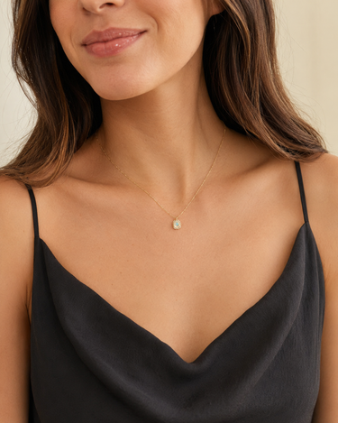 A close-up of a woman wearing a delicate gold chain necklace with a small, round, iridescent white pendant, styled with a black camisole top against a soft beige background.