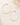 A pair of thin gold hoop earrings with small ball studs, displayed on a light beige textured surface with dried floral accents.