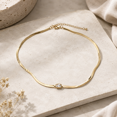 A gold wavy choker necklace with a small clear crystal accent, displayed on a textured beige stone slab with neutral fabric and dried flowers in the background.