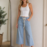 Woman wearing a white tank top and blue jeans standing in a room with a mirror and plant.