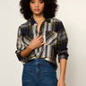 A woman with curly dark hair wearing a long-sleeve plaid button-up shirt in navy, gray, and white, paired with blue jeans, against a plain light background.