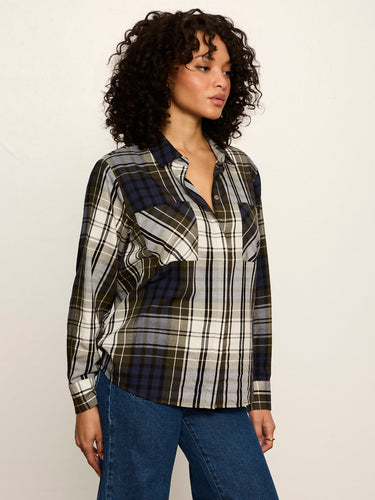 A woman wearing a long-sleeve plaid button-up shirt in black, white, and navy, paired with blue jeans, photographed against a plain off-white studio background.