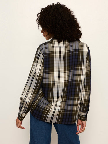 A woman with curly dark hair is shown from the back wearing a long-sleeve plaid button-up shirt in black, white, and blue, paired with blue jeans, against a plain off-white background.