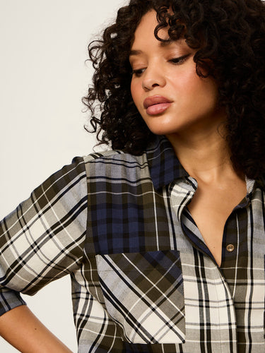A close-up of a woman wearing a plaid button-up shirt with mixed pattern panels in black, white, gray, and navy blue, set against a plain light background.