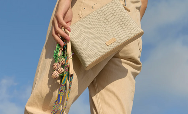 A person wearing beige pants holds a textured beige shoulder bag with a colorful patterned strap against a blue sky background.
