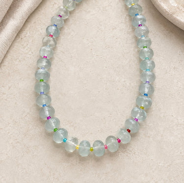 A beaded necklace made of translucent pale blue-green round beads separated by tiny multicolored spacer beads, arranged on a light beige textured surface.