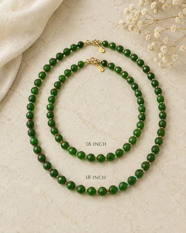 Two green beaded necklaces with gold clasps arranged on a light beige textured surface, with size labels reading 16 INCH and 18 INCH. Decorative fabric and small white flowers appear in the corners.