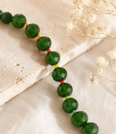 Faceted green gemstone beads strung on a beaded necklace or bracelet, with small accent beads in orange, yellow, red, and light blue, styled on a textured beige stone slab with dried white flowers.