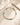 A pastel gradient beaded necklace with green, yellow, and pink round beads, accented by a gold chain clasp, displayed on a textured beige stone surface with soft fabric and stone props.