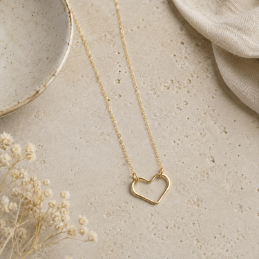 A delicate gold open heart pendant necklace on a fine chain, styled on a textured beige stone surface with dried flowers and neutral fabric props.