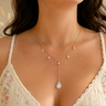 A close-up of a woman wearing a delicate gold lariat necklace with small pearl-like drops and a larger teardrop pendant, styled with a cream floral lace camisole.
