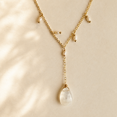 A delicate gold Y-necklace featuring small pearl-like beads and a large teardrop-shaped clear stone pendant, displayed on a warm beige background with soft leaf shadows.