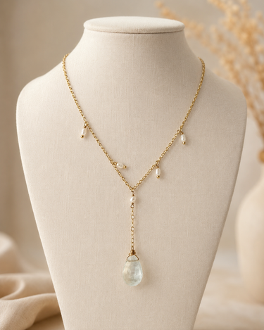 A delicate gold lariat-style necklace with small pearl-like drops and a teardrop-shaped light blue gemstone pendant, displayed on a cream fabric mannequin bust.
