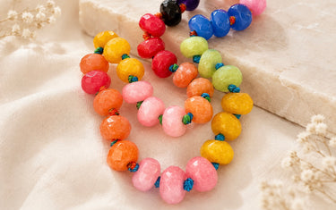 Colorful beaded necklaces arranged on a soft cream fabric with a stone slab in the background. The necklaces feature faceted beads in pink, orange, yellow, green, red, blue, and black, strung on blue cords.