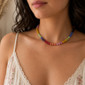 A close-up of a woman wearing a colorful beaded necklace with rainbow-colored beads, paired with a white lace camisole top in a softly lit indoor setting.