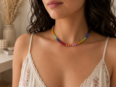 A close-up of a woman wearing a colorful beaded necklace with rainbow-colored beads, paired with a white lace camisole top in a softly lit indoor setting.