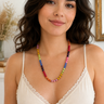 A woman with dark wavy hair wearing a cream lace-trim camisole top and a colorful beaded necklace, photographed in a softly lit indoor setting.