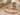 A colorful beaded necklace with gold-toned chain and clasp, arranged in an oval on a light beige textured surface with decorative props including a book, small bowls, and a glass vase.