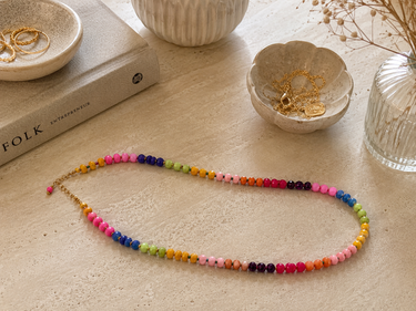 A colorful beaded necklace with gold-toned chain and clasp, arranged in an oval on a light beige textured surface with decorative props including a book, small bowls, and a glass vase.