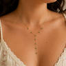 A delicate gold Y-necklace with small green gemstone accents worn by a model in a cream lace-trimmed camisole.