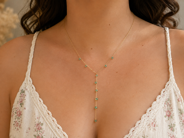 A close-up of a model wearing a delicate gold Y-necklace adorned with small green gemstones, paired with a white floral lace camisole.