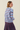 A woman shown from the back wearing a light blue knit sweater with horizontal navy and red stripes, paired with a pleated skirt, against a solid light beige background.
