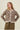 A woman modeling a fuzzy leopard-print cardigan with brown buttons, paired with olive green pants, against a solid light beige background.