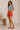 Woman wearing a black and white striped shirt and orange shorts standing in a minimalistic room.