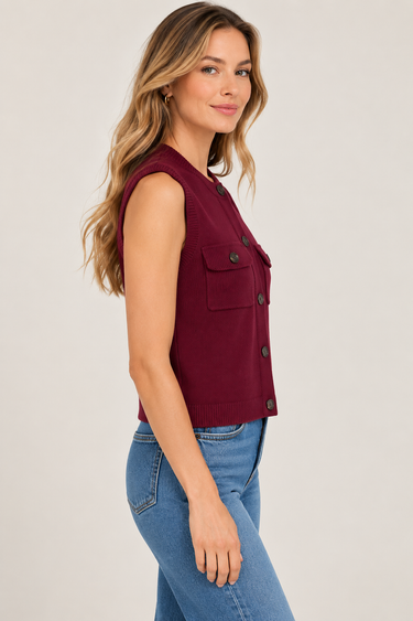 A woman wearing a sleeveless burgundy knit vest with button details and a chest pocket, paired with blue jeans, photographed against a light gray studio background.